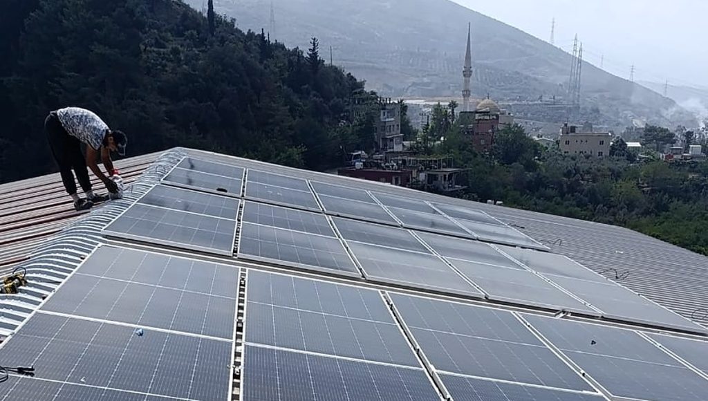 Says Enerji ekibinden bir kişi çatıda panel montajı yaparken, arkada Hatay yamaçları